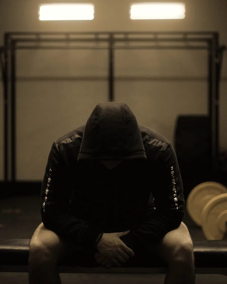 A hooded athlete sitting head down on a bench in a dim gym