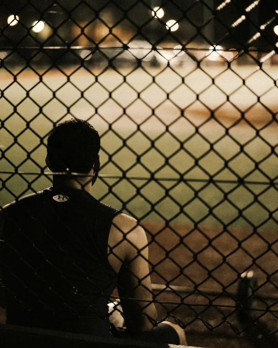 An athlete sitting alone behind the backstop, watching a floodlit field at night