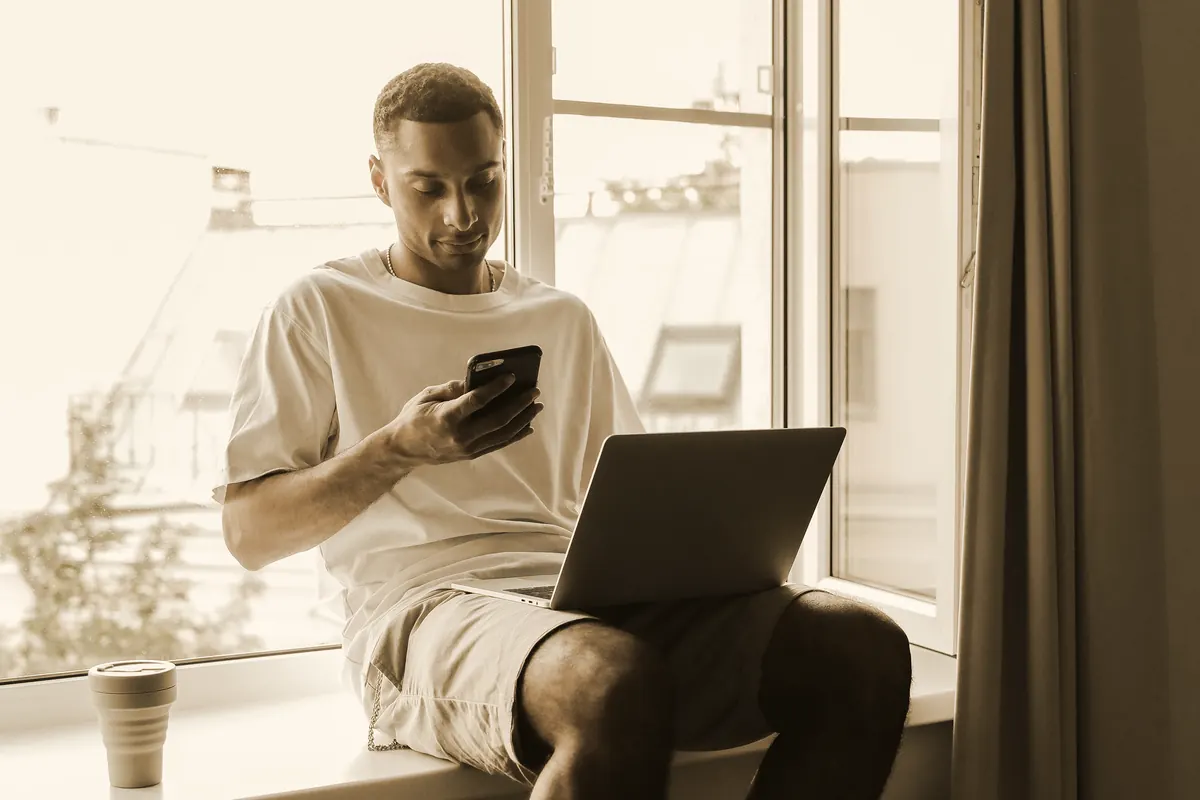 A young man sitting on a windowsill with a laptop open on his lap, looking at his phone instead